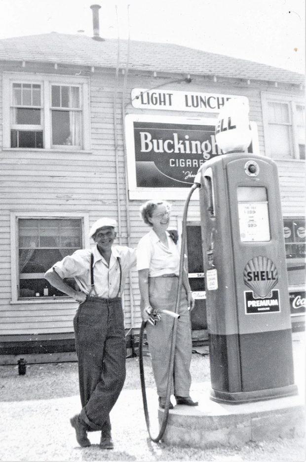 Photo en noir et blanc de deux personnes devant une pompe à essence, avec un grand édifice en bois à l’arrière-plan
