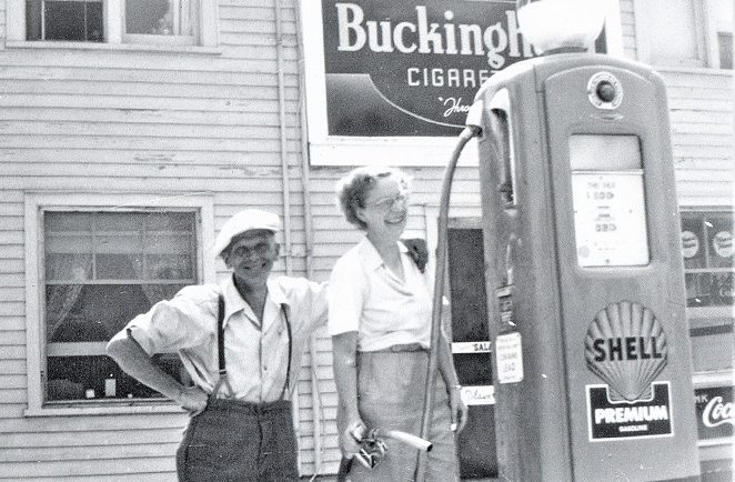 Photo en noir et blanc de deux personnes devant une pompe à essence, avec un grand édifice en bois à l’arrière-plan