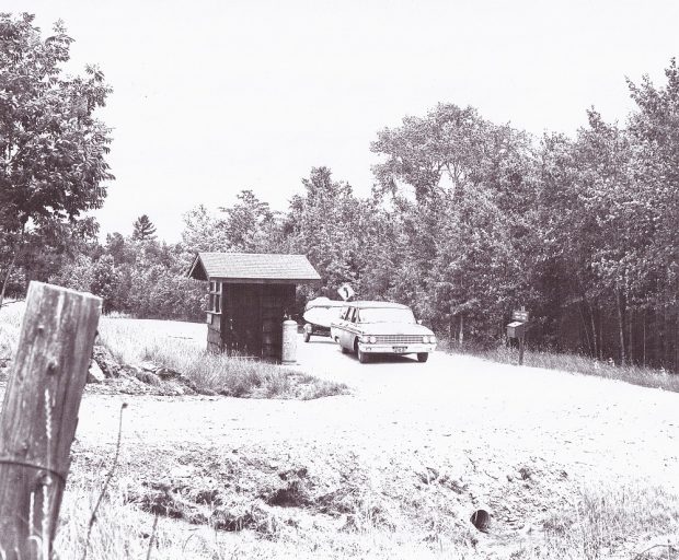 Photo en noir et blanc d’un véhicule ancien qui remorque un bateau sur une route de terre près d’une cabane, avec des arbres à l'arrière-plan