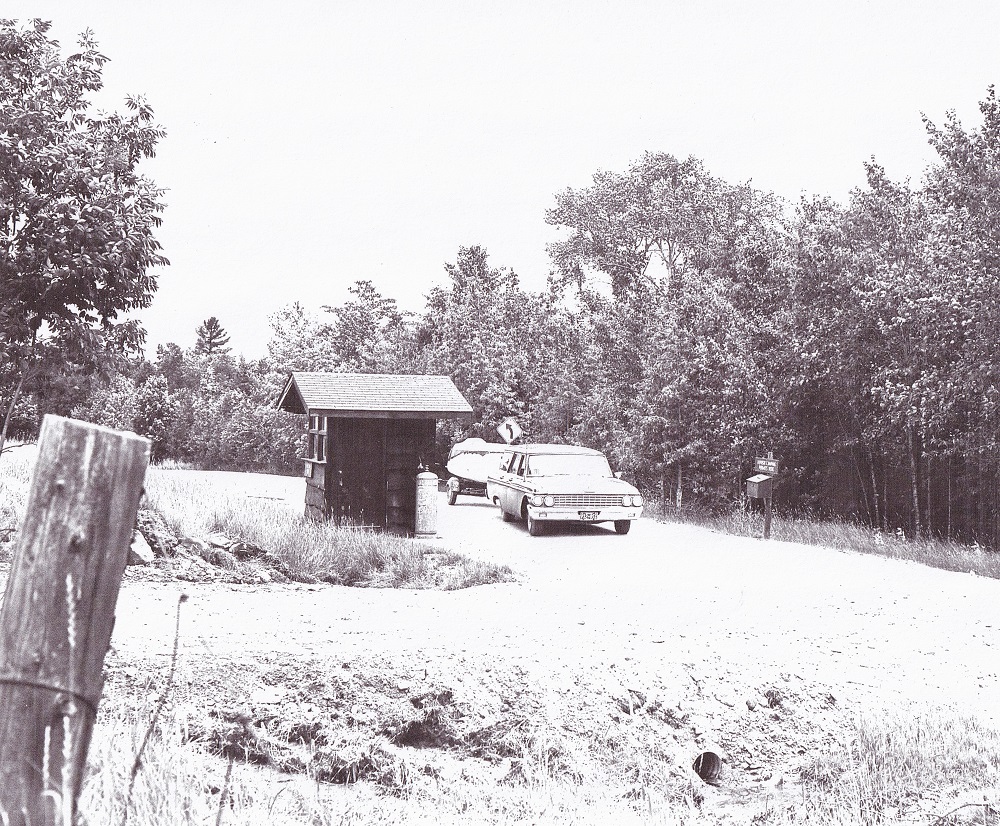 Photo en noir et blanc d’un véhicule ancien qui remorque un bateau sur une route de terre près d’une cabane, avec des arbres à l'arrière-plan