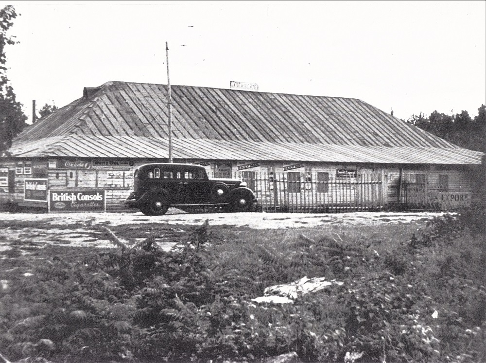Photo en noir et blanc d’une voiture ancienne stationnée devant un bâtiment avec un large toit en bois