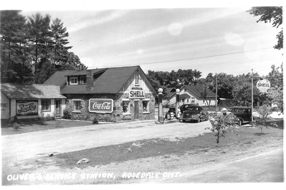 Photo en noir et blanc de voitures anciennes stationnées devant les pompes à essence avec l’édifice en pierre à l’arrière-plan