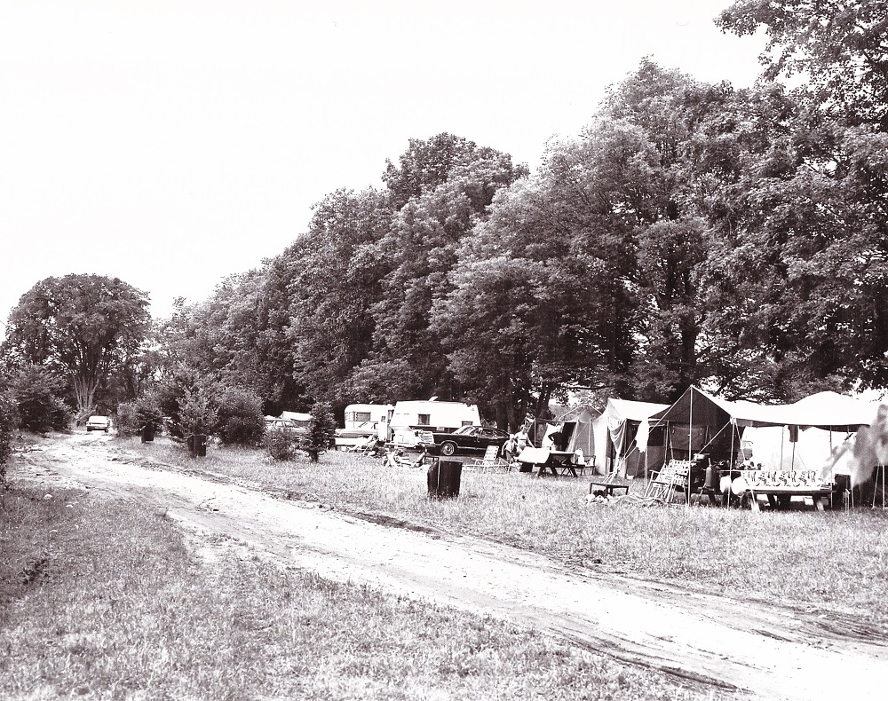 Photo en noir et blanc de tentes et roulottes stationnées sous des arbres, entourées d’équipement de camping, avec un chemin boueux à l'avant-plan