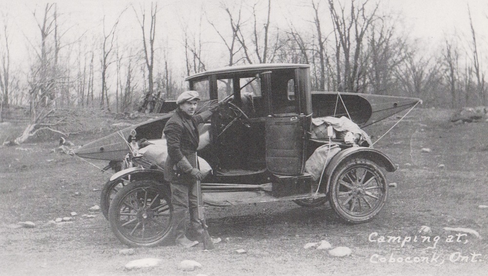 Photo en noir et blanc d’un homme à côté d’un véhicule ancien qui remorque un canoë et de l’équipement de camping, avec des arbres à l'arrière-plan