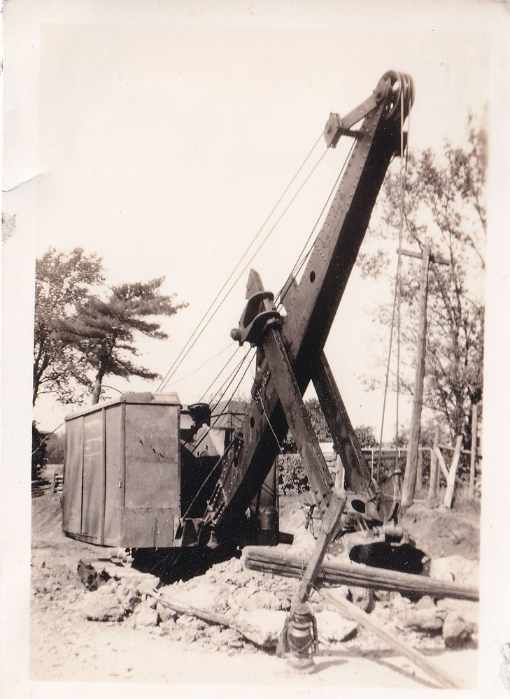 Photo en noir et blanc d’une pelle de vapeur qui creuse au bord de la route, avec des arbres et des poteaux électriques à l'arrière-plan