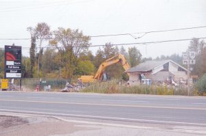 Photo couleur d’une machine qui démolit un édifice, avec des arbres à l'arrière-plan, ainsi qu’une affiche et une autoroute à l'avant-plan