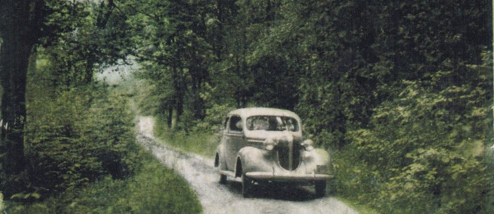 Photo couleur d’un véhicule ancien qui longe une rue bordée par des arbres et du feuillage