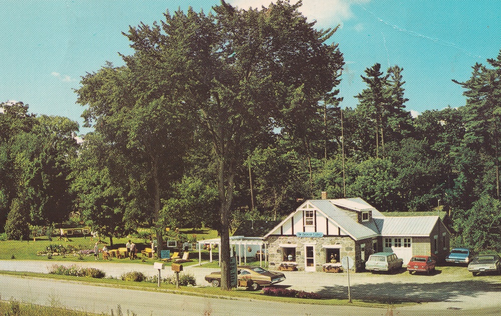 Photo couleur d’un édifice de pierre entouré par des jardins et des arbres, avec des voitures et un parc de stationnement à l'avant-plan