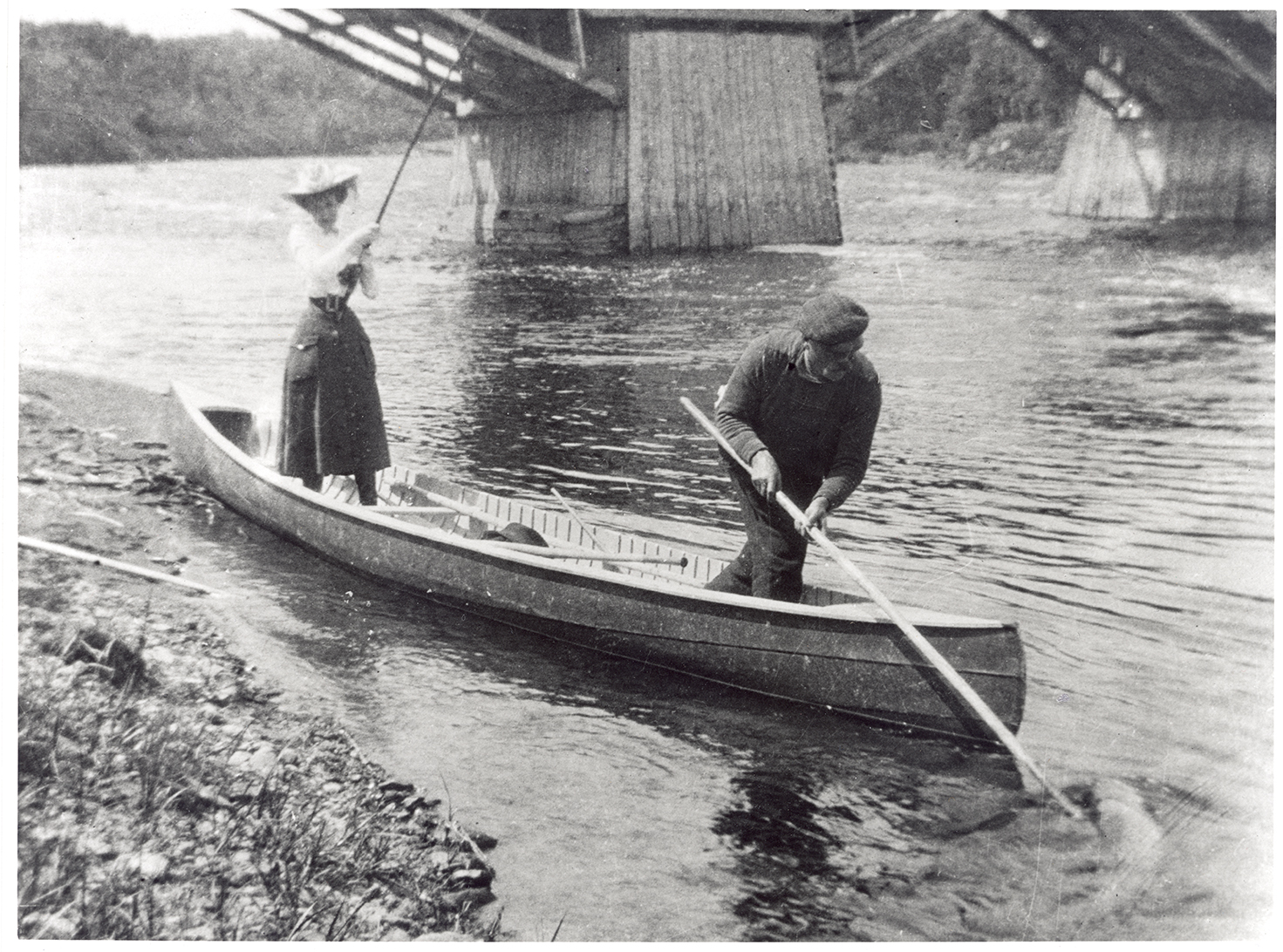 Épreuve argentique montrant Elsie Reford, debout dans un canot sur la berge de la rivière. Elle est vêtue d'une jupe mi-longue, une chemise blanche et d'un chapeau élégant. Elle semble tirer une longue canne à pêche. Un homme, situé au devant du canot sort un gros poissons de la rivière a l'aide d'une longue pôle et d'un crochet.