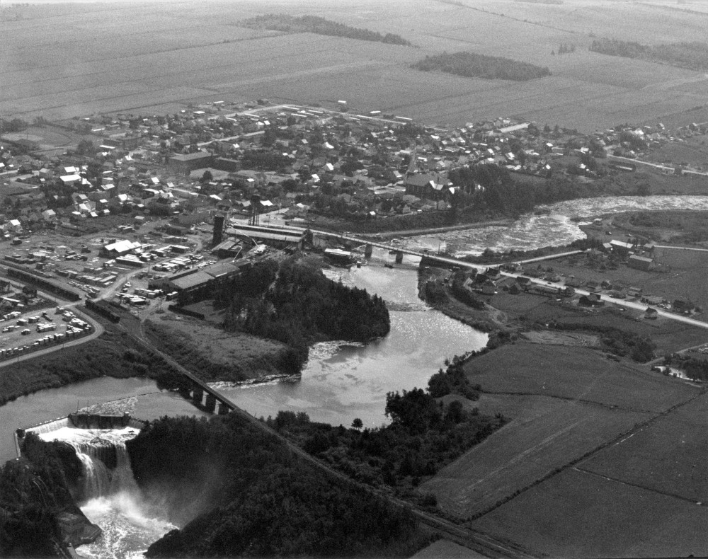 Cette photographie aérienne montre le village de Price au pic de sa prospérité. La compagnie de la Price Brothers occupait un large territoire voisin de la rivière. On retrouve aussi de chaque côté de la rivière les terminale de chemin de fer du Canada et du Golf.