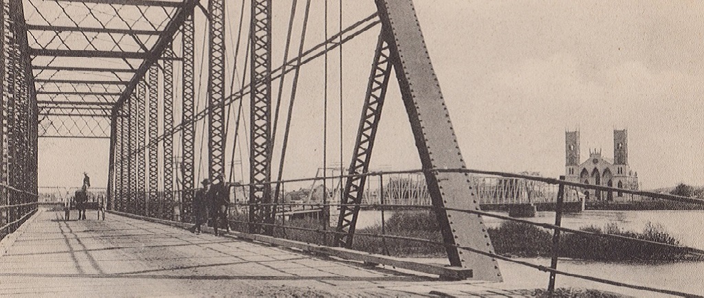 Vue du pont de Sainte-Anne-de-la-Pérade enjambant la rivière Sainte-Anne et sur lequel se trouvent une voiture à chevaux conduite par deux hommes ainsi que deux autres hommes appuyés sur la rambarde. Au loin on aperçoit l’Église Sainte-Anne.