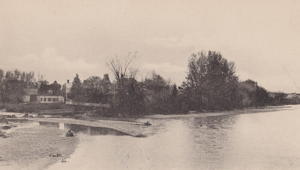 Photographie en noir et blanc de la maison de John Jones Ross sur l’Île Saint-Ignace vue depuis le pont municipal et dont le toit dépasse des arbres.