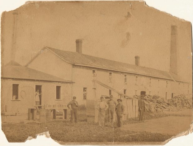 Photographie d’archives du bâtiment abritant la manufacture d’allumettes devant laquelle se trouvent plusieurs travailleurs. Des piles de débris de bois longent la façade percée de nombreuses fenêtres où apparaissent des travailleurs et travailleuses.