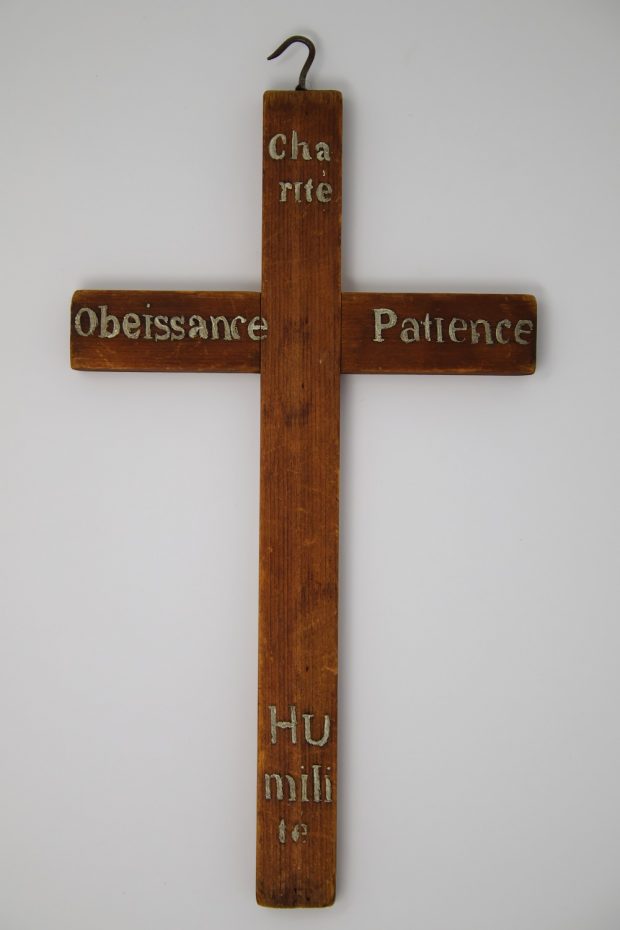 Photographie d'une croix de bois brun aux extrémités de laquelle apparaissent les mots Charité Obéissance Patience et Humilité inscrits en lettres dorées.