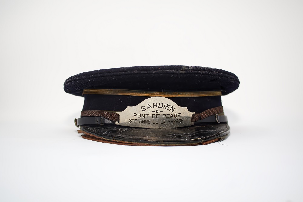 Casquette noire avec lacet doré vue de face où apparaît l’inscription Gardien Pont de péage Sainte Anne de la Pérade gravée sur un écusson en métal.