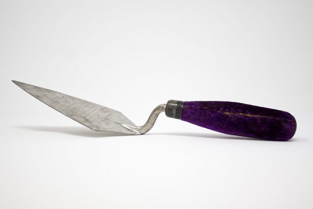 Truelle en métal avec manche recouvert de velours violet sur laquelle est gravée une inscription.