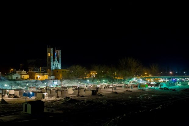 Photographie nocturne des dizaines de cabanes de pêcheurs sur la rivière Sainte-Anne devant l’église illuminée.