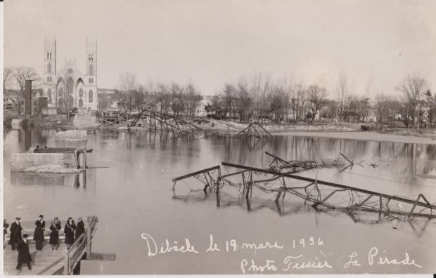 Photographie en noir et blanc du pont de fer détruit par la débâcle du 19 mars 1936 avec les restes de la structure dans la rivière et l’église Sainte-Anne sur l’une des rives. Un groupe de sept personnes constate les dégâts à partir d’un bout restant de la passerelle sur l’autre rive.