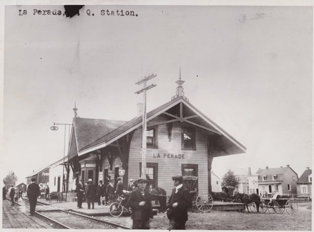 Photographie en noir et blanc de la gare de Sainte-Anne-de-la-Pérade où s’attroupent des voyageurs et voitures à chevaux.