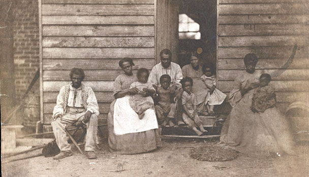 Photos d’un groupe d’esclaves devant la case qui leur sert de logis.  