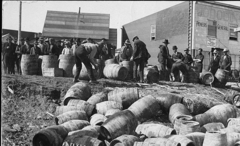 Des ouvriers éventrent des barils pour laisser s’échapper l’alcool qu’ils contiennent; les tonneaux vides s’entassent au pied d’une pente.