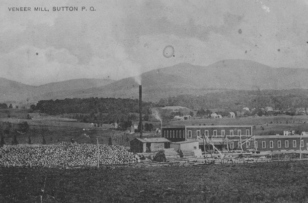 En avant-plan, l’usine de contreplaqué de la Veneer Mill sur la rue Western à Sutton et sa cour à bois remplie de billots; en arrière-plan , les monts Sutton.