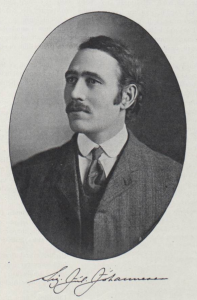 Portrait photographique ovale en noir et blanc d’un homme, cadré à partir des épaules. Il a les cheveux noirs, coiffés sur le côté, avec des favoris et une moustache. Vêtu d’un costume et d’une cravate, il regarde vers la gauche. Ci-dessous la photo, sa signature cursive est imprimée.