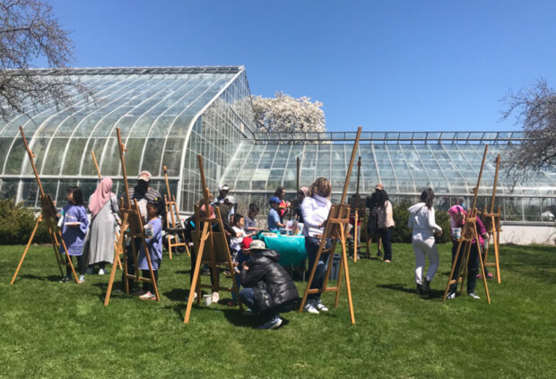 Une photographie d'un atelier de peinture en plein air avec des enfants et des adultes qui peignent sur des chevalets dans un parc. La scène est ensoleillée avec une serre en arrière-plan.