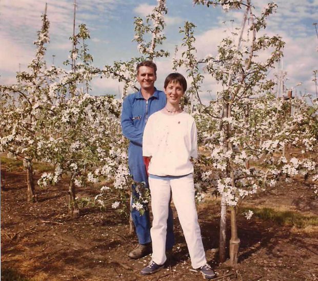 Photo couleur qui présente une femme, Diane Goyette et un homme, Louis Poulin dans leur verger. La femme est vêtue de blanc tandis que l’homme est en bleu. Derrière eux, on peut voir des pommiers nains.