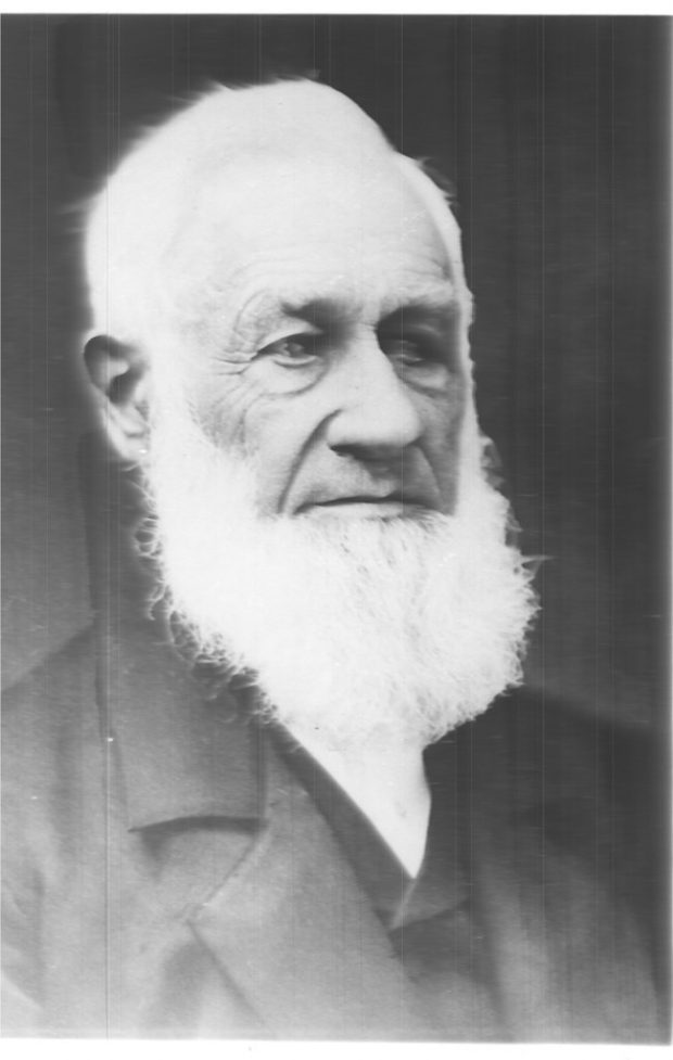 Photo noir et blanc qui présente le portrait de biais de Aaron Alexander Adams portant un costume noir avec des cheveux blancs et une barbe blanche.