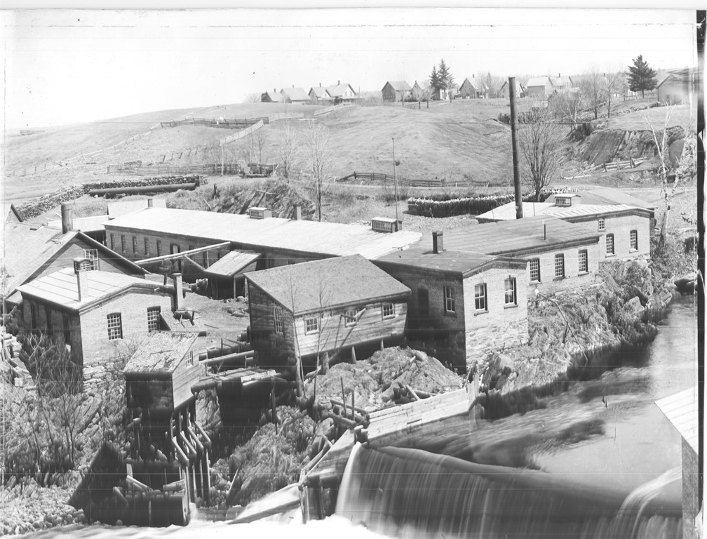 Photo noir et blanc d’un paysage avec plusieurs bâtiments d’une usine avec une chute de la rivière Coaticook en avant-plan. Au loin, on voit une colline et des habitations. 