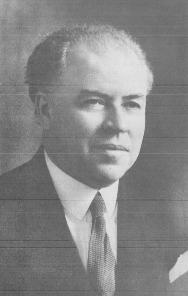 Photo noir et blanc présentant le portrait de biais du Dr Ernest Gendreau. Il a les cheveux gris, porte un costume sombre, une chemise blanche, une pochette blanche ainsi qu’une cravate blanc-gris.  