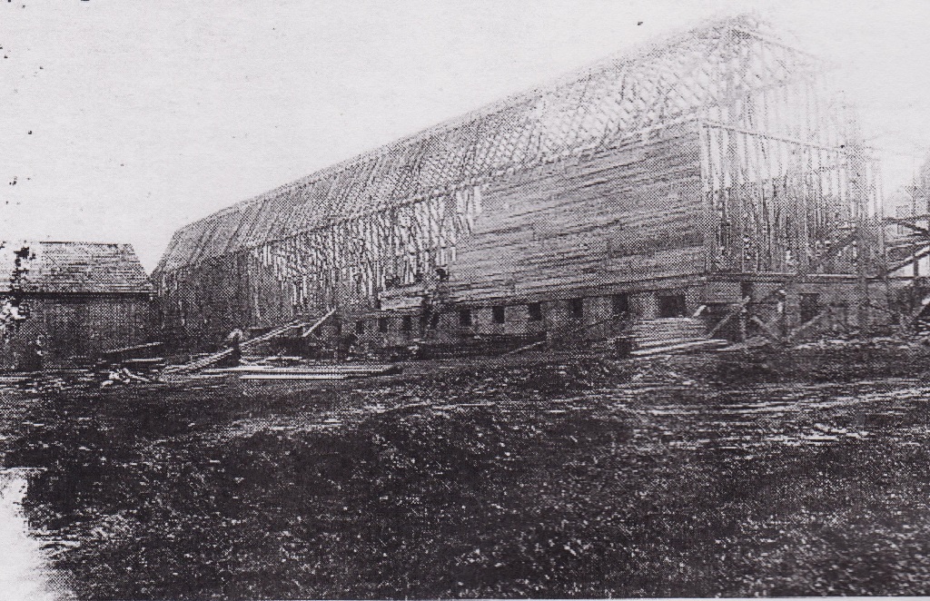 Photographie noir et blanc d’une énorme structure en construction dont la charpente est érigée. Des ouvriers commencent à la recouvrir de planches.