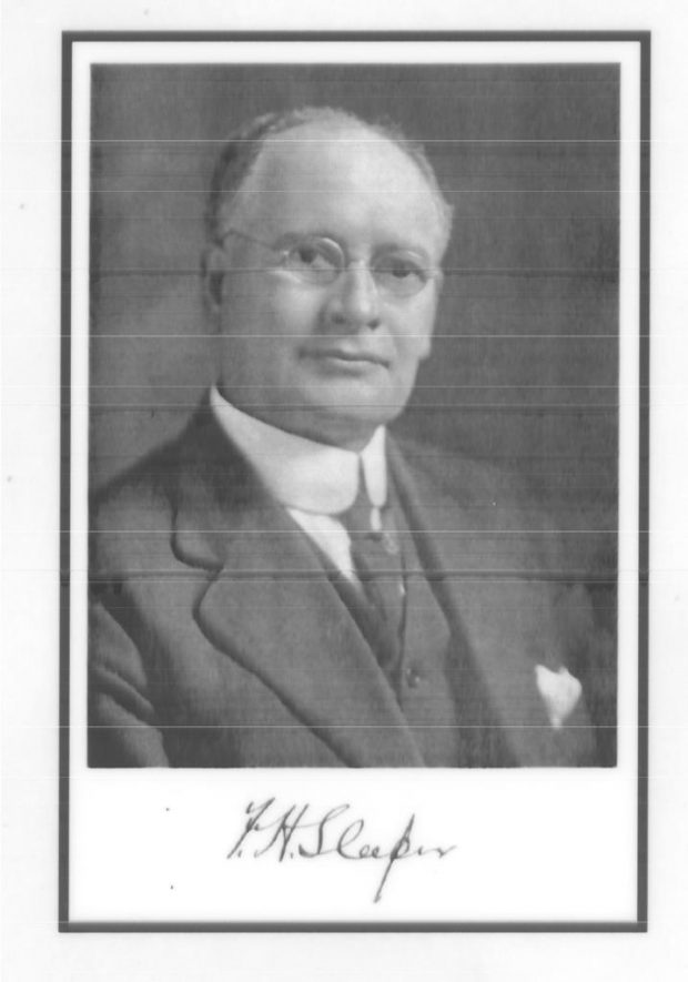 Photo noir et blanc présentant le portrait de biais de Frank Sleeper qui porte des lunettes, un costume et une cravate sombres ainsi qu’une chemise et une pochette blanches.