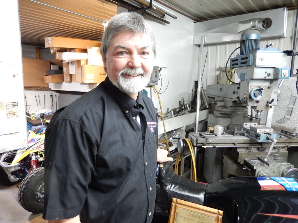 Photo couleur qui présente le portrait de l’inventeur Gilles Gagné en chemise noire, légèrement en biais avec des cheveux gris, une moustache et une barbe blanche. En arrière-plan on a une vue partielle de son atelier avec des machines, des outils et du matériel de rangement.