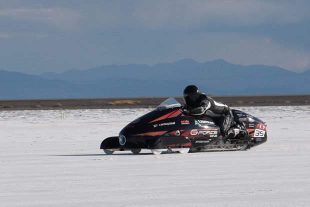 Photo couleur qui présente un engin monté par une personne sur une piste de course. On peut voir une étendue de neige, une étendue de terre ferme puis, à l’horizon, une chaîne de montagnes.