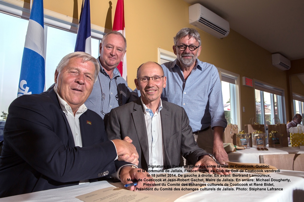 Photo couleur d’un groupe de personnes : assis, deux hommes vêtus de costumes sombres et de chemises blanches se serrent la main; deux autres portant des chemises bleues sont debout. Derrière eux se trouvent, de gauche à droite, le drapeau du Québec, le drapeau de la France ainsi que celui du Canada. On peut également apercevoir des cadeaux emballés placés sur des tables.