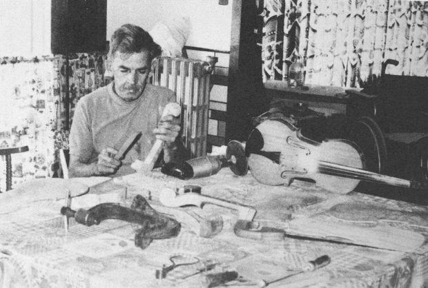 Photo noir et blanc qui présente un homme assis chez lui, sur une table, habillé en tee-shirt gris, en train de travailler. Sur la table se trouvent un violon, des morceaux de bois et des outils.