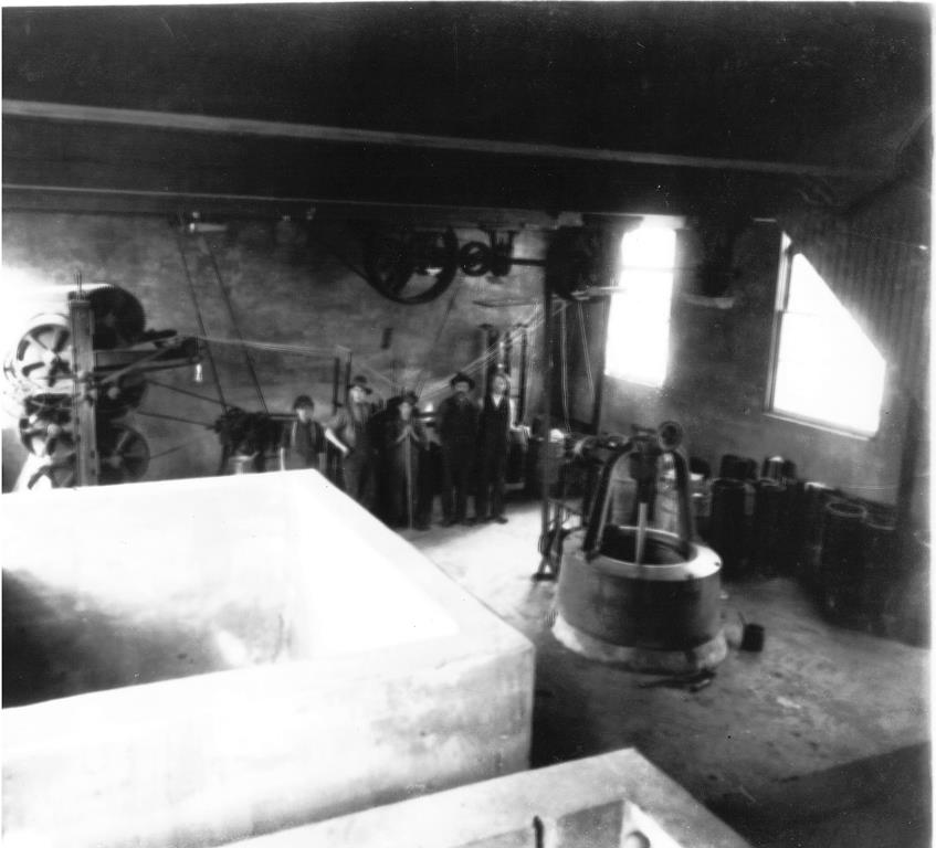 Photo noir et blanc de l’intérieur d’une usine avec des installations, du matériel, des outils et des ouvriers. 