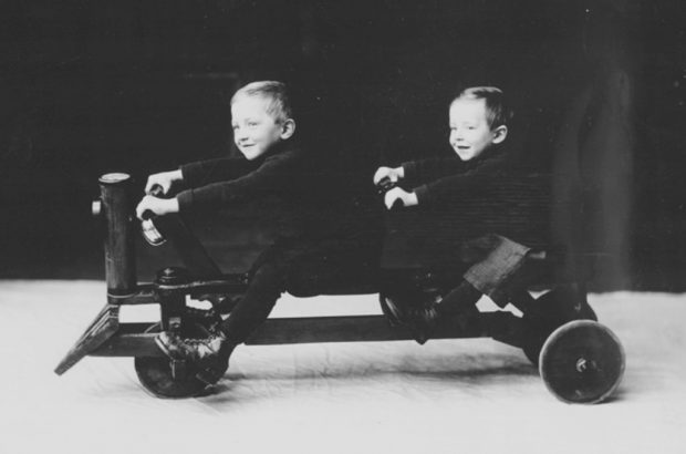 Photo ancienne noir et blanc avec 2 petits enfants souriants assis sur une voiturette à 3 roues avec 2 guidons. 