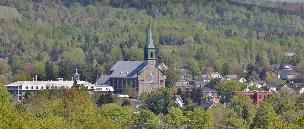 Photo couleur d’une vue partielle de Coaticook dans un paysage boisé. On peut voir l’Église St-Edmond avec son clocher, le grand bâtiment du Collège Rivier et d’autres bâtisses.