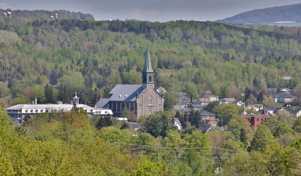 Photo couleur d’une vue partielle de Coaticook dans un paysage boisé. On peut voir l’Église St-Edmond avec son clocher, le grand bâtiment du Collège Rivier et d’autres bâtisses.