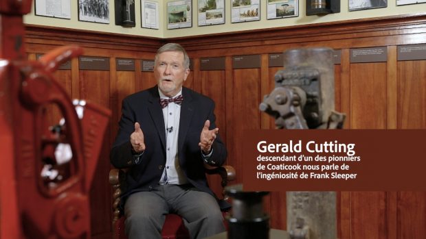 Photo couleur présentant Gerald Cutting, descendant d’un des pionniers de Coaticook, vêtu d’un pantalon gris, d’une chemise bleu clair, d’un nœud papillon rouge-gris et d’un veston noir. Il est assis dans une salle d’exposition du Musée Beaulne et se trouve entre deux crics.