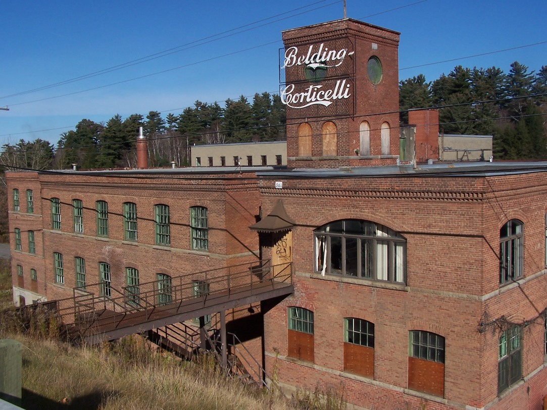 Photo couleur du bâtiment de l’usine Belding Corticelli, construit en brique rouge, compte 4 étages et est surmontée d’un belvédère sur lequel est inscrit le nom de l’usine en lettres stylisées. Une passerelle relie le bâtiment au chemin.