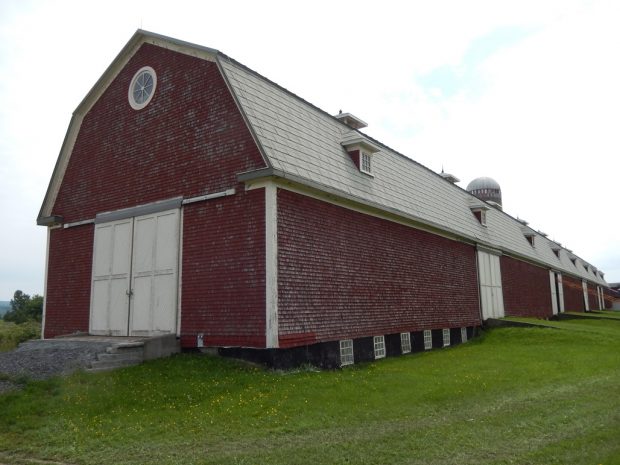 Photo couleur de la grange de la Ferme-du-Plateau dans toute sa longueur. Le bâtiment est de couleur bourgogne avec des portes blanches.