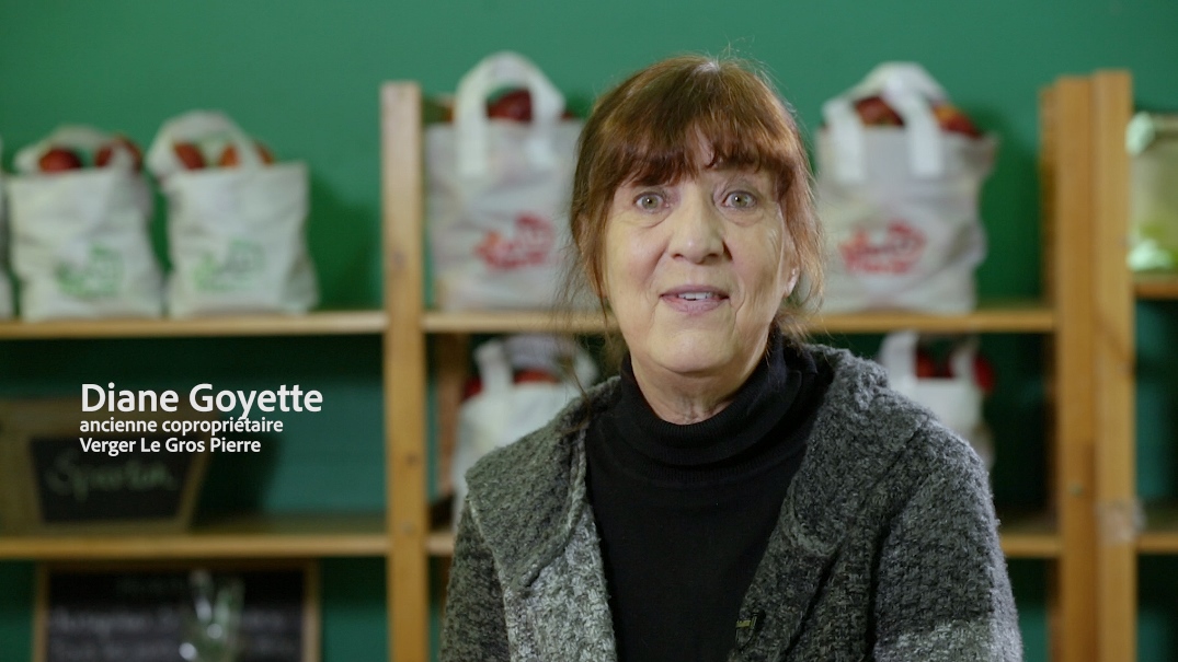 Photo couleur présentant Diane Goyette, ancienne copropriétaire du Verger Le Gros Pierre, vêtue d’un pull noir et d’un manteau gris. Derrière elle se trouve une étagère sur laquelle sont disposés des sacs de pommes.