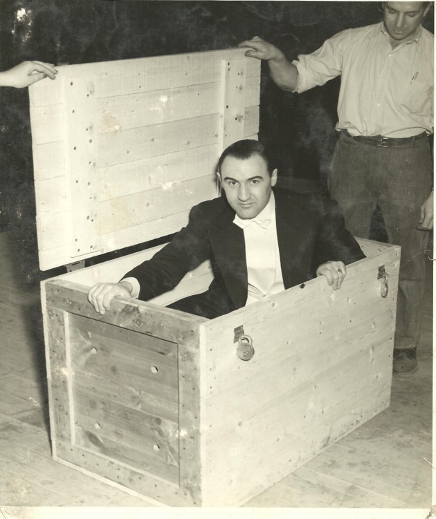 Photo noir et blanc qui présente le Grand Henri assis dans une caisse en bois, vêtu d’un costume noir, d’une chemise blanche et d’un nœud papillon blanc. Debout à droite se trouve un homme vêtu d’un pantalon sombre et d’une chemise claire et qui tient le couvercle de la caisse. En haut à gauche, on peut voir une main qui tient également le couvercle de la caisse.