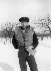 Dick Uyede debout dehors en hiver avec des arbres derrière lui portant un gilet de fourrure