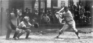 Un homme prêt à frapper une balle de baseball avec l'arbitre et le receveur derrière lui et un groupe de personnes regardant au-delà d'une clôture.