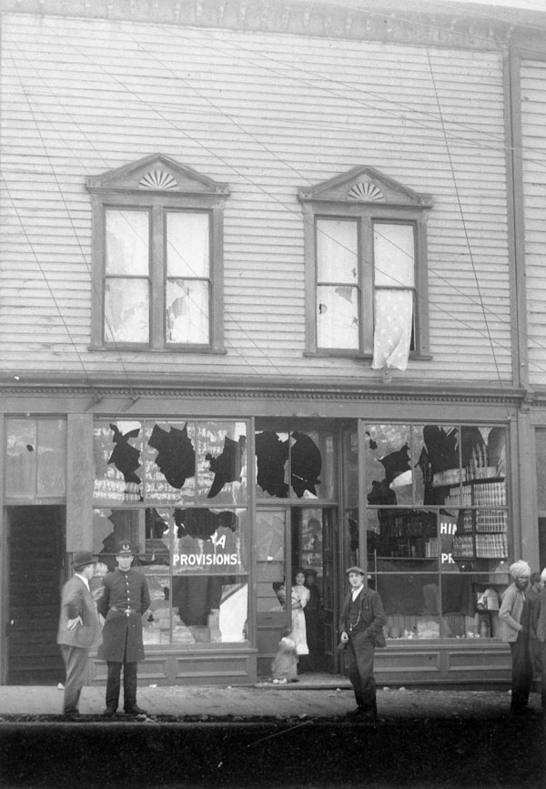 Des vitres brisées dans un commerce avec la police à l'extérieur et une femme japonaise dans l'embrasure de la porte.